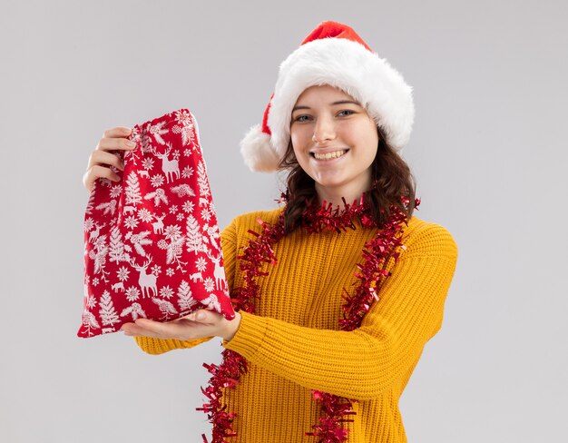 Smiling young slavic girl with santa hat and with garland around neck holds christmas gift bag isolated on white wall with copy space