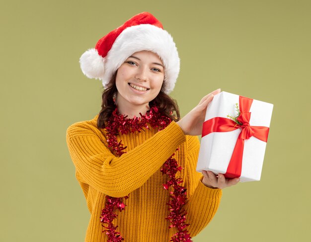 Smiling young slavic girl with santa hat and with garland around neck holds christmas gift box isolated on olive green wall with copy space