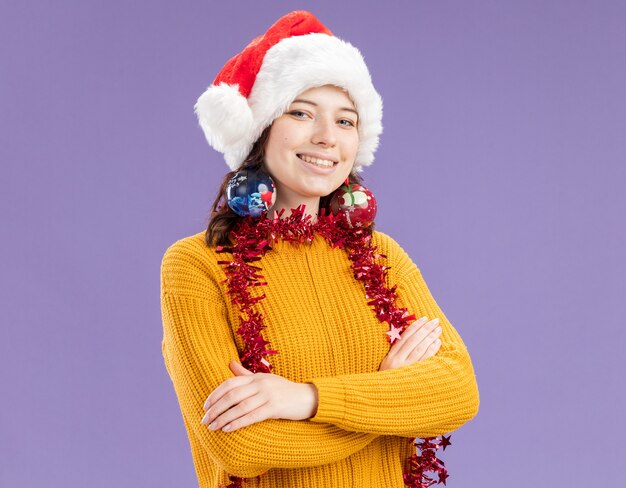 Smiling young slavic girl with santa hat and with garland around neck holds glass ball ornaments on ears standing with crossed arms isolated on purple wall with copy space