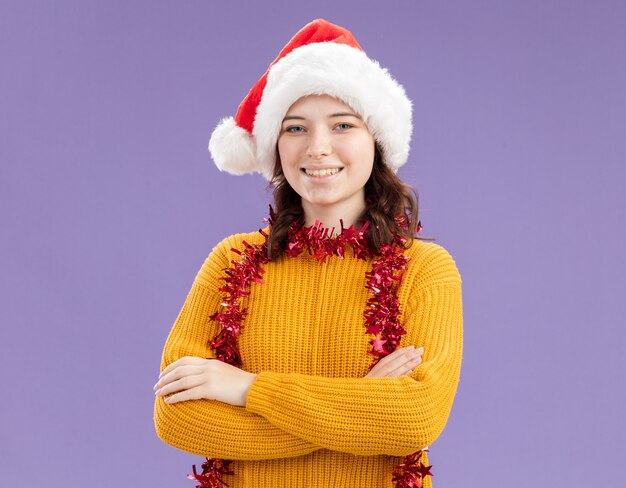 Smiling young slavic girl with santa hat and with garland around neck standing with crossed arms isolated on purple background with copy space