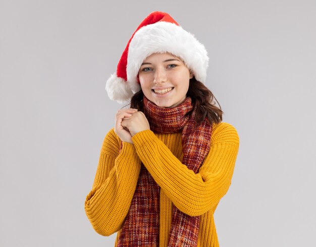 Smiling young slavic girl with santa hat and with scarf around neck holds hands together and looks 
