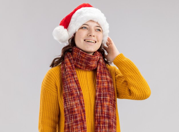Улыбающаяся молодая славянская девушка в новогодней шапке и с шарфом на шее кладет руку на лицо и смотрит на сторону, изолированную на белой стене с копией пространства