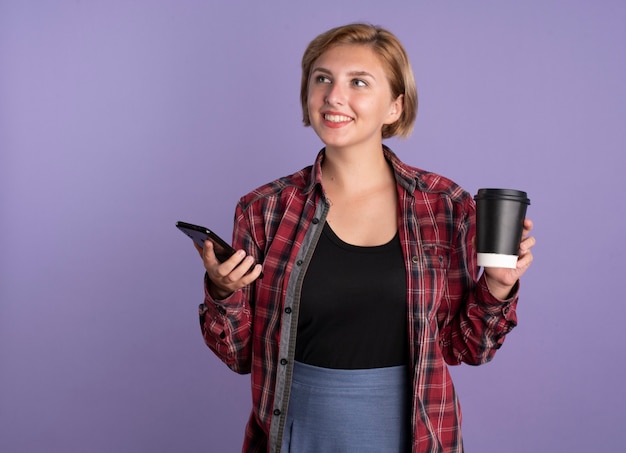 Улыбающаяся молодая славянская студентка держит телефон и чашку, глядя в сторону