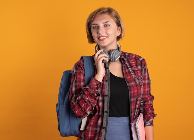 Smiling young slavic student girl wearing backpack holds headphones around neck 