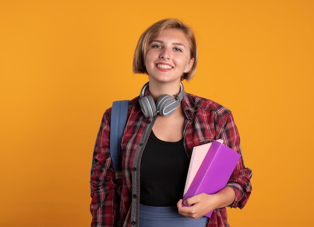 Smiling young slavic student girl with headphones wearing backpack holding book and notebook