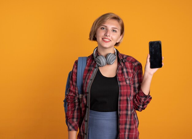 携帯電話を保持しているバックパックを着てヘッドフォンで笑顔の若いスラブ学生の女の子