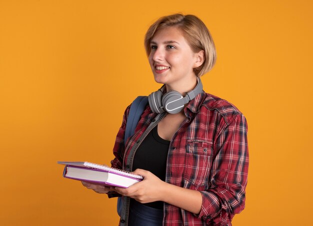 バックパックを着たヘッドフォンで笑顔の若いスラブ学生の女の子が本とノートを持っている