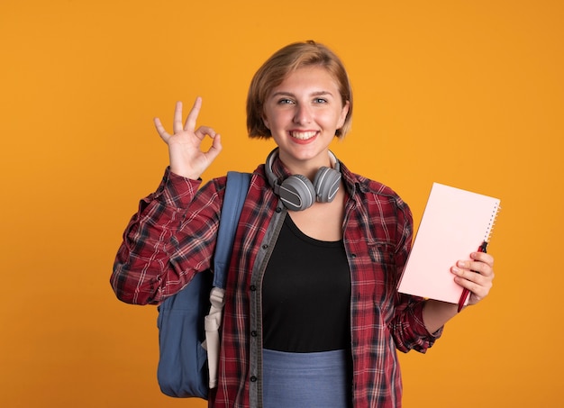 バックパックを着たヘッドフォンを付けた笑顔の若いスラブ学生の女の子は、ノートとペンのジェスチャー、OK ハンドサインを保持します