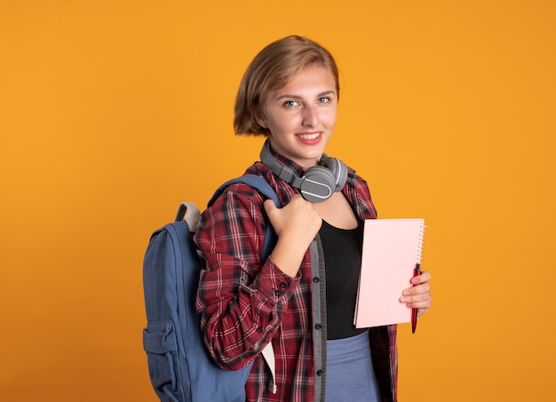 배낭을 착용하는 헤드폰으로 웃는 젊은 슬라브 학생 소녀가 카메라를보고 노트북과 펜을 보유하고 있습니다.