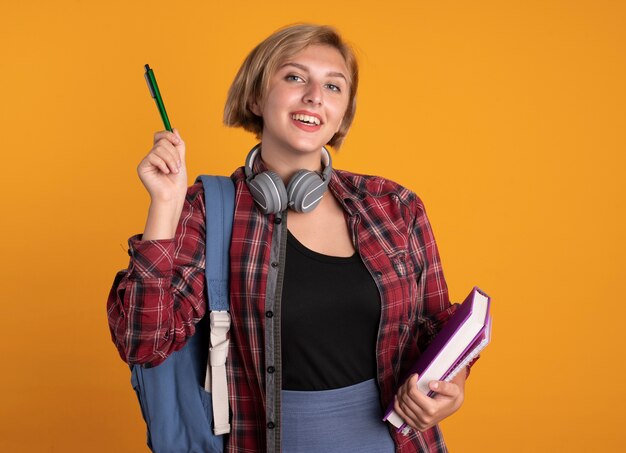 배낭을 착용하는 헤드폰으로 웃는 젊은 슬라브 학생 소녀는 펜 책과 노트북을 보유하고 있습니다.
