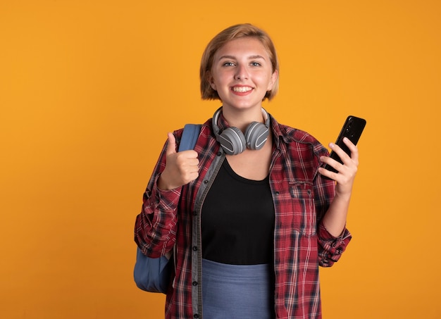 배낭을 착용하는 헤드폰으로 웃는 젊은 슬라브 학생 소녀는 전화와 엄지 손가락을 보유하고 있습니다.