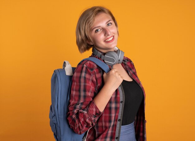 Smiling young slavic student girl with headphones wearing backpack stands sideways 