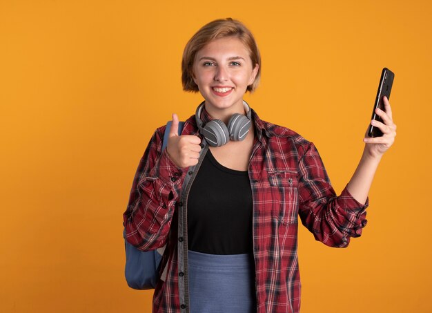 Улыбающаяся молодая славянская студентка с наушниками в рюкзаке показывает палец вверх и держит телефон