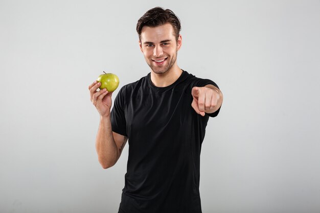 Улыбающийся молодой спортивный человек держит яблоко, указывая на вас.