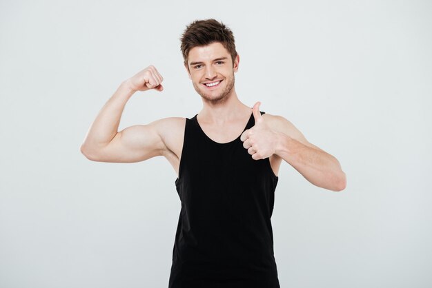 Smiling young sportsman flexing biceps and showing thumbs up gesture