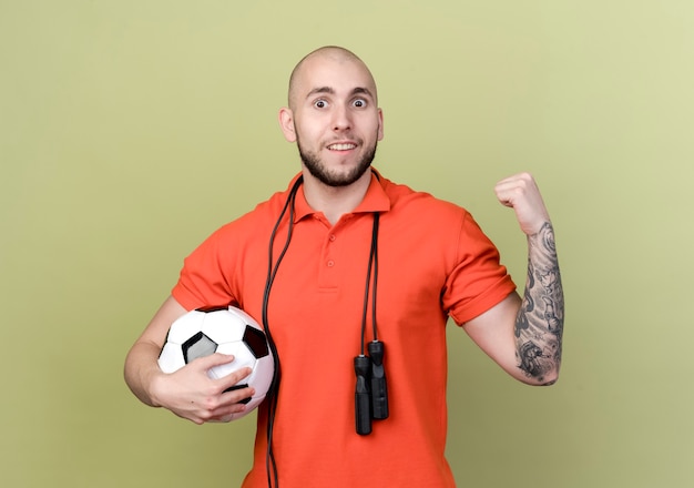 Free photo smiling young sporty man holding ball with jump rope on shoulder and showing yes gesture isolated on olive green wall