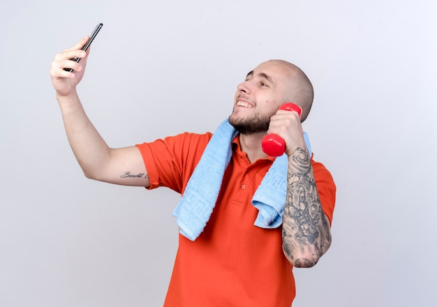 Smiling young sporty man holding dumbbell with towel on shoulder and take a selfie isolated on white