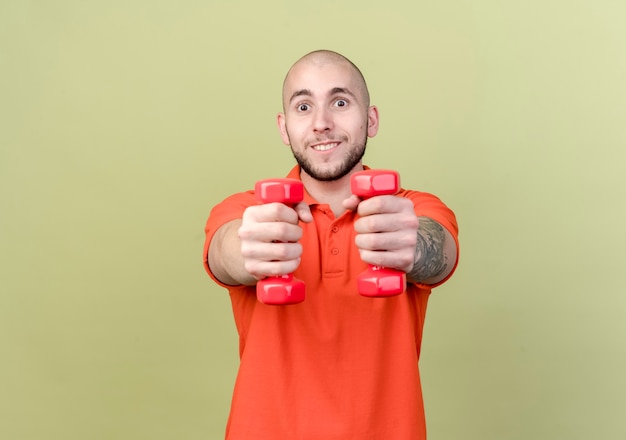 Smiling young sporty man holding out dumbbells isolated on olive green wall