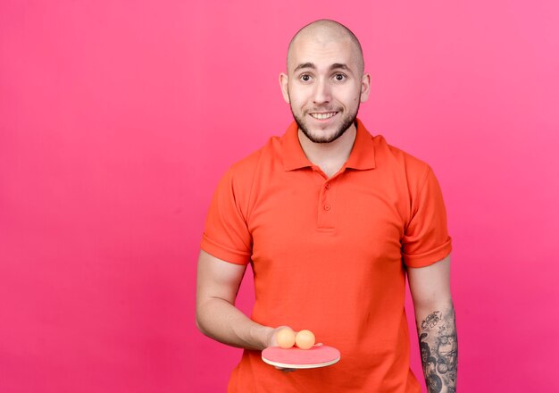 Smiling young sporty man holding ping pong racket with balls isolated on pink wall