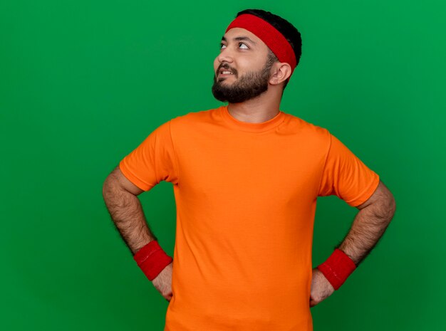 Smiling young sporty man looking at side wearing headband and wristband putting hands on hip isolated on green background