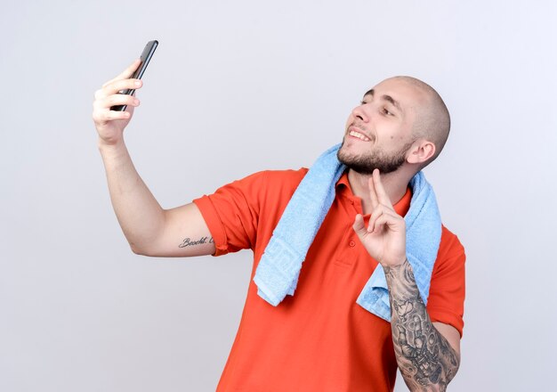 Smiling young sporty man showing peace gesture and take a selfie isolated on white wall