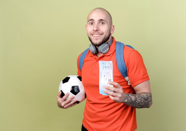 Улыбающийся молодой спортивный мужчина в задней сумке и наушниках на шее держит мяч с билетами, изолированным на оливково-зеленой стене с копией пространства