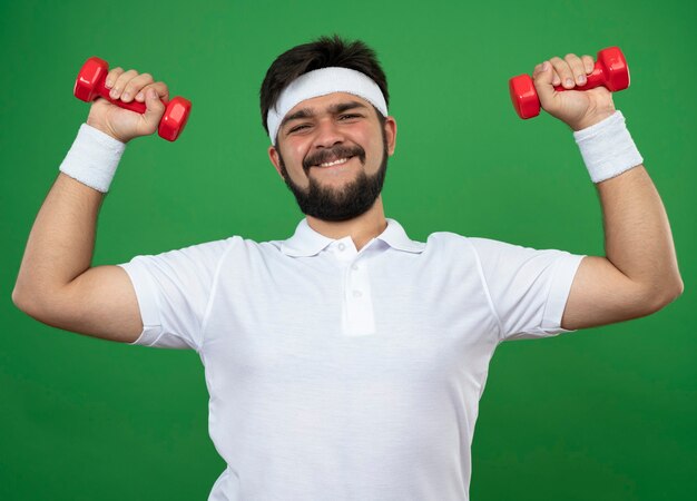 Улыбающийся молодой спортивный мужчина с повязкой на голову и браслетом, тренирующимся с гантелями, изолированными на зеленом