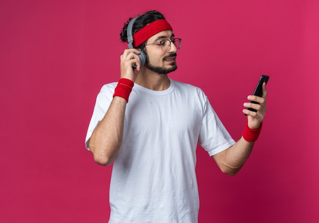 Улыбающийся молодой спортивный человек с повязкой на голову с браслетом и наушниками, держащий и смотрящий на телефон
