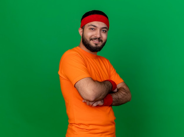 Smiling young sporty man wearing headband and wristband crossing hands isolated on green background