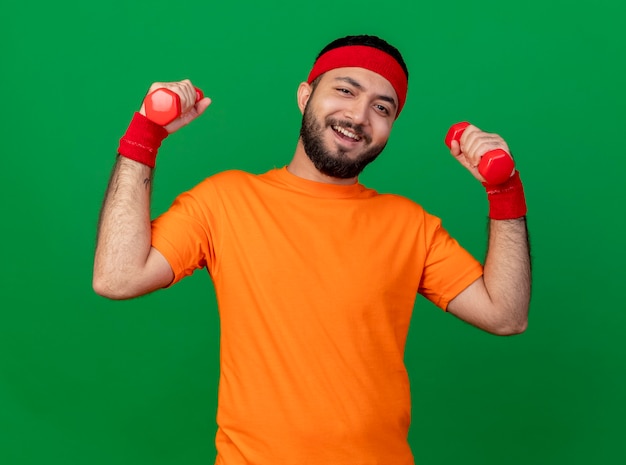 Smiling young sporty man wearing headband and wristband exercising with dumbbells isolated on green background