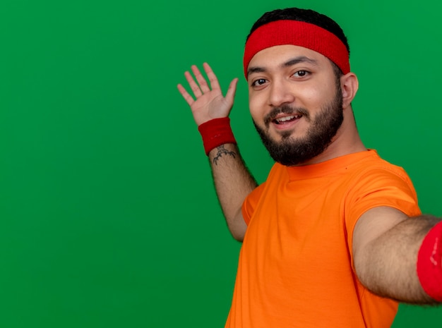 Free photo smiling young sporty man wearing headband and wristband holding camera points