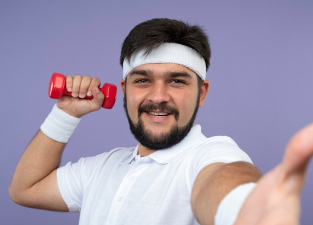 Smiling young sporty man wearing headband and wristband holding dumbbell isolated on green wall