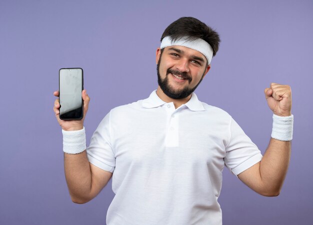Улыбающийся молодой спортивный мужчина с повязкой на голову и браслетом, держащим телефон и показывающим жест `` да ''