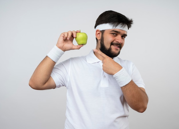 Улыбающийся молодой спортивный мужчина с повязкой на голову и браслетом держит и указывает на яблоко, изолированное на белой стене