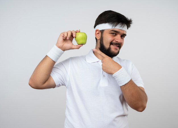 Smiling young sporty man wearing headband and wristband holding and points at apple isolated on white wall