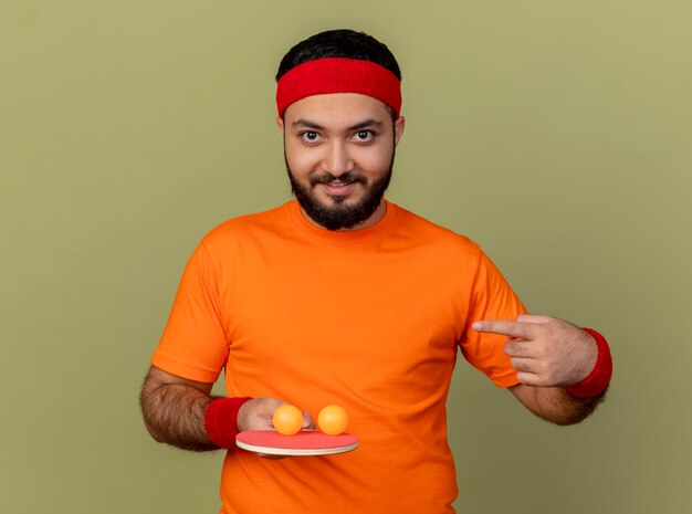 Smiling young sporty man wearing headband and wristband holding and points at ping pong racket with balls isolated on olive green background