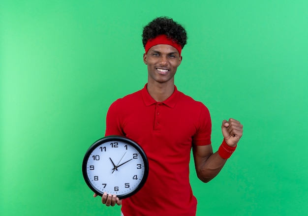 Smiling young sporty man wearing headband and wristband holding wall clock