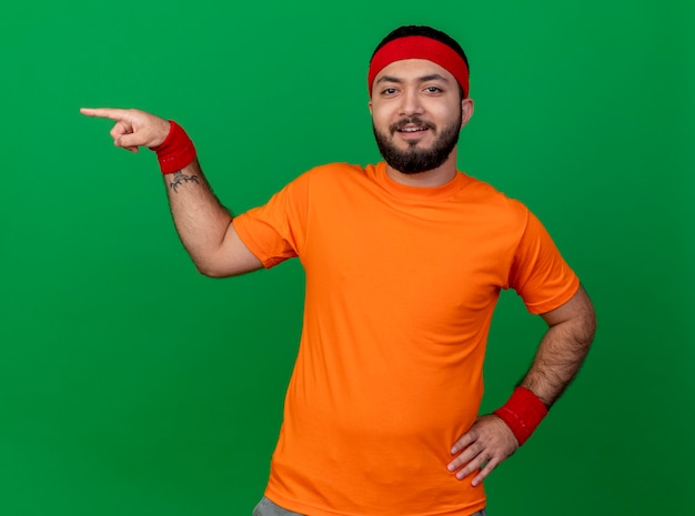 Smiling young sporty man wearing headband and wristband points at side and putting hand on hip isolated on green background with copy space