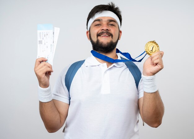 Smiling young sporty man wearing headband and wristband with backpack holding tickets with medal isolated on white wall