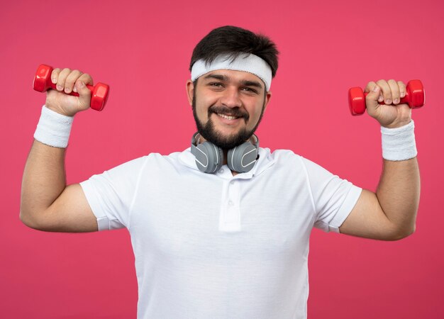 Smiling young sporty man wearing headband and wristband with headphones exercising with dumbbells