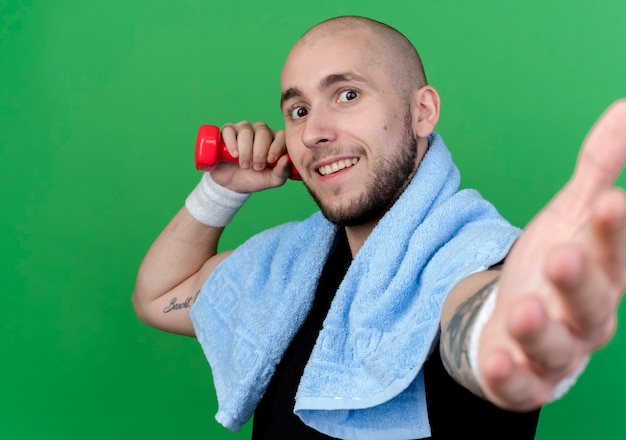 Free photo smiling young sporty man wearing wristband holding dumbbell with towel on shoulder and holding out hand isolated on green wall