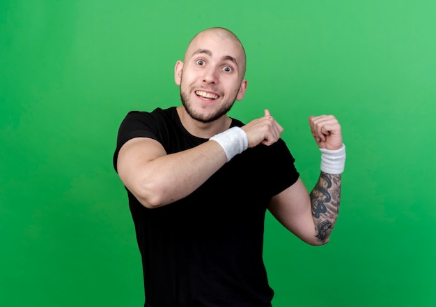 Smiling young sporty man wearing wristband points at back isolated on green