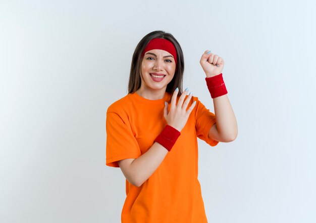Smiling young sporty woman wearing headband and wristbands clenching fist looking keeping hand in air isolated