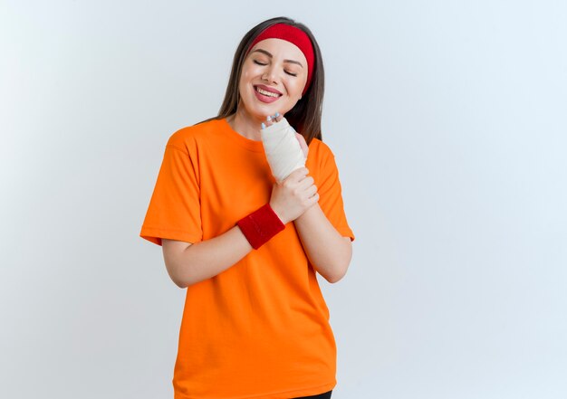 Smiling young sporty woman wearing headband and wristbands holding injured wrist wrapped in bandage with closed eyes isolated on white wall with copy space