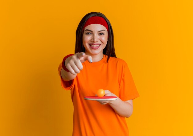 ヘッドバンドとリストバンドを身に着けている笑顔の若いスポーティな女性は、その上にボールが付いているピンポンラケットを持って、コピースペースのあるオレンジ色の壁に孤立して見ていると指している