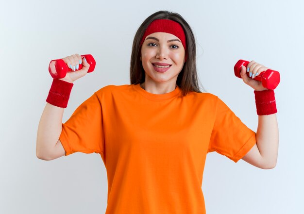 Smiling young sporty woman wearing headband and wristbands looking holding dumbbells 
