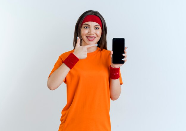 Sorridente giovane donna sportiva che indossa la fascia e braccialetti che mostrano il telefono cellulare che punta a guardare isolato