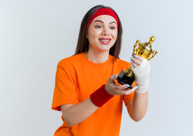 Smiling young sporty woman wearing headband and wristbands with injured wrist wrapped in bandage holding and looking at winner cup isolated on white wall with copy space