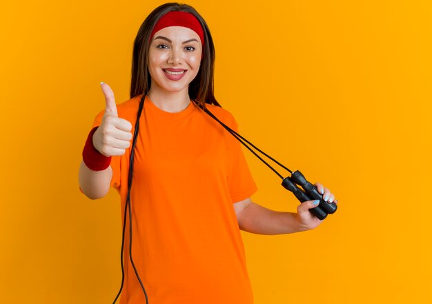 Smiling young sporty woman wearing headband and wristbands with jump rope around neck grabbing jump rope looking showing thumb up 