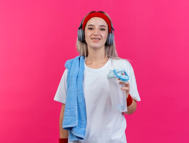 Smiling young sporty woman with braces on headphones wearing headband and wristbands holds water bottle and towel on shoulder isolated on pink wall